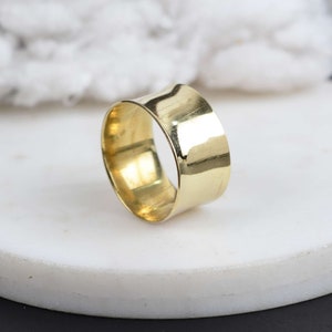 Può includere: Un anello a fascia larga in tonalità oro lucido con una superficie liscia e riflettente. L'anello presenta un design circolare semplice e continuo con una larghezza uniforme, mostrando una finitura metallica brillante che cattura la luce.
