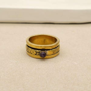 Könnte beinhalten: Ein goldfarbener Ring mit floralem Design und einem kleinen, runden, lila Edelstein. Der Ring hat ein mehrschichtiges Design mit geätzten floralen Mustern. Der Edelstein ist in der Mitte des Rings gefasst.