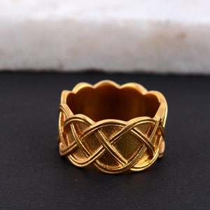 Könnte beinhalten: Ein goldfarbener Ring mit einem geflochtenen Design. Der Ring hat eine strukturierte Oberfläche und einen gewellten Innenrand. Das komplizierte Muster erzeugt einen auffälligen Look. Der Ring wird auf einer schwarzen Oberfläche präsentiert.