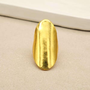 Könnte beinhalten: Ein goldfarbener Ring mit einer großen, länglichen, ovalen Vorderseite und einer glatten, leicht gewölbten Oberfläche. Das Metall hat ein poliertes Finish mit subtiler Textur, was ein reflektierendes Erscheinungsbild entlang der Mittelachse erzeugt.