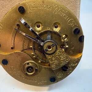 May include: Close-up of a vintage Illinois Watch Co. pocket watch movement. The gold-toned mechanism features intricate gears, hands, and decorative engravings. The text "Illinois Watch Co." and "Springfield, Ill." are visible, along with the serial number 414877.