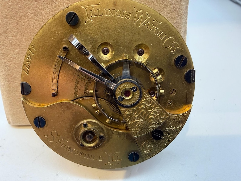 May include: Close-up of a vintage Illinois Watch Co. pocket watch movement. The gold-toned mechanism features intricate gears, hands, and decorative engravings. The text "Illinois Watch Co." and "Springfield, Ill." are visible, along with the serial number 414877.