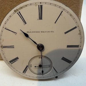 May include: Antique watch face with black Roman numerals and hands on a white background. The words "ILLINOIS WATCH CO." are printed above the center. A smaller seconds dial is at the bottom. The watch face is slightly worn.