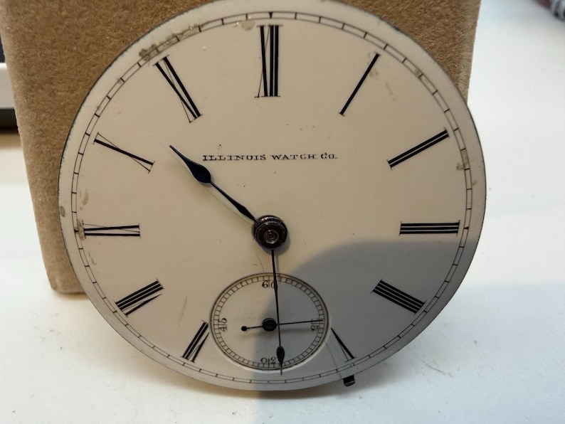 May include: Antique watch face with black Roman numerals and hands on a white background. The words "ILLINOIS WATCH CO." are printed above the center. A smaller seconds dial is at the bottom. The watch face is slightly worn.