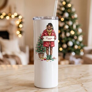 May include: A white stainless steel tumbler with a clear lid and straw, featuring an illustration of a person in a red puffer jacket, yellow scarf, and green boots holding a sign that reads Denise 2026 next to a small Christmas tree.