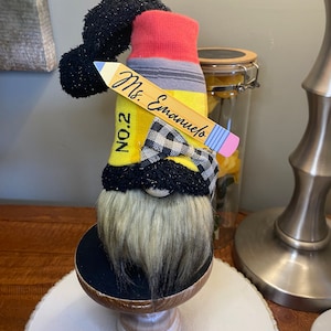 Può includere: Un gnomo a forma di matita gialla con un papillon bianco e nero, un cappello rosso e grigio e un'etichetta di legno con il nome "Ms. Emmanuel" e "No. 2".