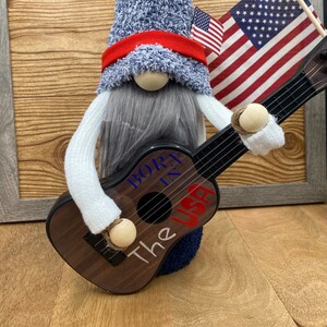 Puede incluir: Un gnomo con un sombrero azul y rojo y pantalones vaqueros azules está tocando una guitarra acústica marrón. La guitarra tiene las palabras "Born in the USA" escritas en ella. El gnomo está de pie frente a un marco de madera con una bandera estadounidense colgada en él.