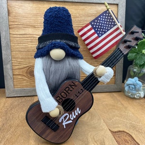 Puede incluir: Un gnomo azul y gris con una larga barba blanca sostiene una guitarra de madera. La guitarra tiene las palabras "Born to Run" escritas en ella. El gnomo lleva un sombrero azul con una banda negra.