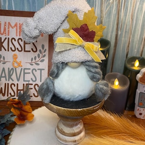 May include: A handmade gnome doll with a gray plush hat and beard, adorned with fall-themed leaves and a yellow ribbon. The gnome sits on a wooden pedestal, with a sign that reads "Pumpkin Kisses & Harvest Wishes" in the background. Decorative candles and fall foliage complete the autumn scene.