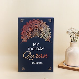 May include: A dark blue journal with a gold and orange mandala design at the top. The text on the cover reads "MY 100-DAY Quran JOURNAL" in white and orange lettering. The journal has a rectangular shape with decorative corner accents.