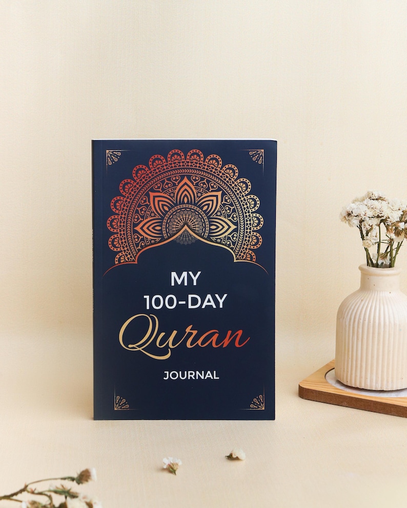 May include: A dark blue journal with a gold and orange mandala design at the top. The text on the cover reads "MY 100-DAY Quran JOURNAL" in white and orange lettering. The journal has a rectangular shape with decorative corner accents.