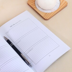 May include: An open journal with white pages featuring printed sections labeled DAY 26: Verse 3:191, Arabic, Translation, and Notes/Reflection. A black and silver ballpoint pen rests diagonally across the page. The page number 29 is visible.