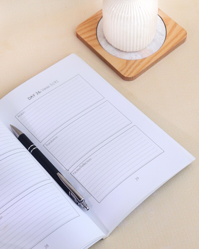May include: An open journal with white pages featuring printed sections labeled DAY 26: Verse 3:191, Arabic, Translation, and Notes/Reflection. A black and silver ballpoint pen rests diagonally across the page. The page number 29 is visible.