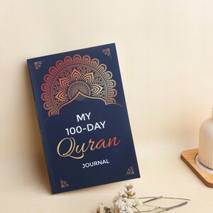 May include: A dark blue journal with a gold and orange gradient mandala design at the top. The cover features the white text "MY 100-DAY" and the orange script text "Quran" followed by the white word "JOURNAL" centered on the front cover.