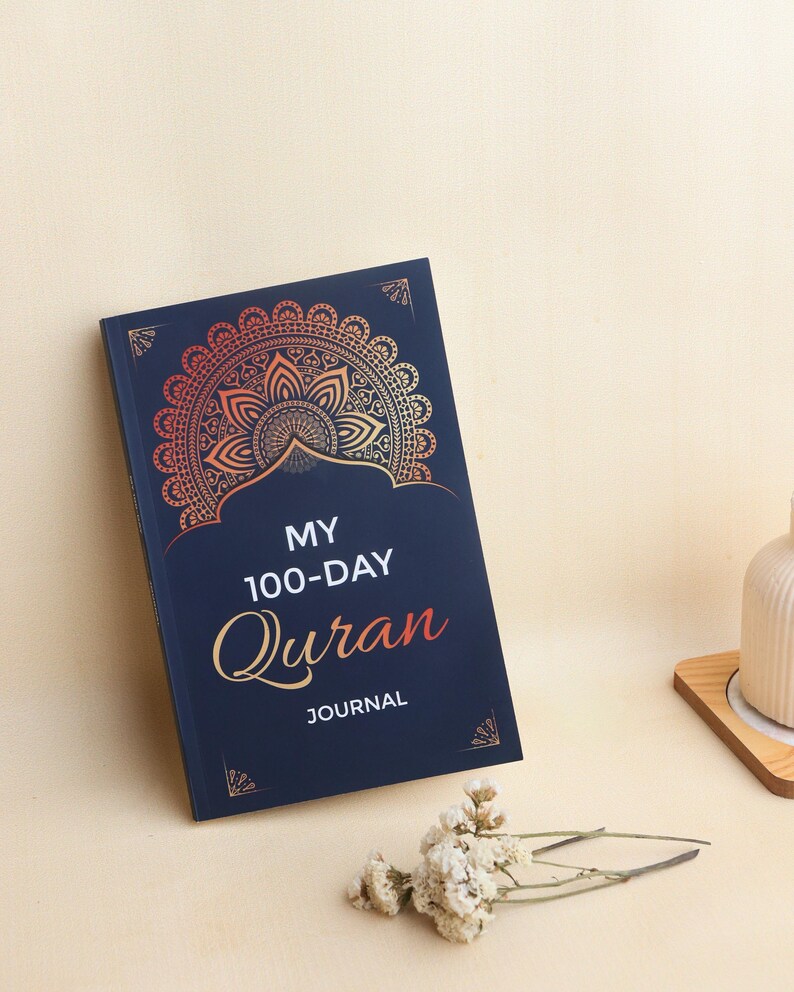 May include: A dark blue journal with a gold and orange gradient mandala design at the top. The cover features the white text "MY 100-DAY" and the orange script text "Quran" followed by the white word "JOURNAL" centered on the front cover.