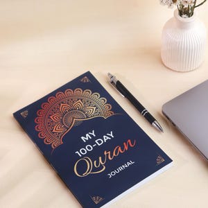 May include: A dark blue journal with a gold and orange mandala design on the cover. The text reads MY 100-DAY QURAN JOURNAL in white and orange lettering. The journal is positioned next to a black pen, a laptop corner, and a small white vase.