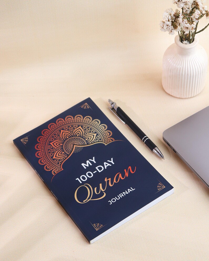 May include: A dark blue journal with a gold and orange mandala design on the cover. The text reads MY 100-DAY QURAN JOURNAL in white and orange lettering. The journal is positioned next to a black pen, a laptop corner, and a small white vase.
