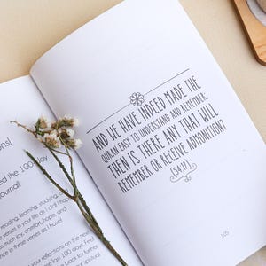 May include: An open book with white pages featuring black printed text that reads, And we have indeed made the Quran easy to understand and remember: then is there any that will remember or receive admonition? (54:17). A dried flower stem lies across.