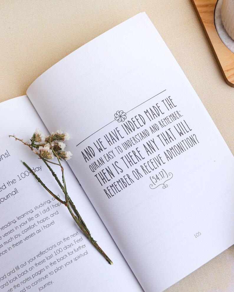 May include: An open book with white pages featuring black printed text that reads, And we have indeed made the Quran easy to understand and remember: then is there any that will remember or receive admonition? (54:17). A dried flower stem lies across.