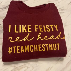 Może przedstawiać: Bordowa bluza z okrągłym dekoltem i złotym napisem I LIKE FEISTY red heads #TEAMCHESTNUT. Tekst jest wyświetlany w połączeniu pogrubionych wielkich liter oraz kursywy, umieszczonych na przedniej części tego elementu garderoby.
