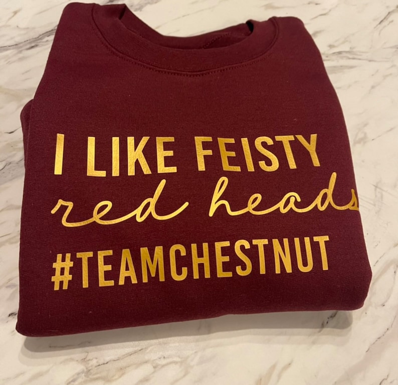 Może przedstawiać: Bordowa bluza z okrągłym dekoltem i złotym napisem I LIKE FEISTY red heads #TEAMCHESTNUT. Tekst jest wyświetlany w połączeniu pogrubionych wielkich liter oraz kursywy, umieszczonych na przedniej części tego elementu garderoby.