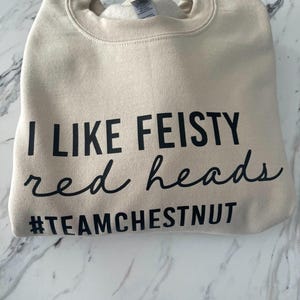 Może przedstawiać: Beżowa bluza z okrągłym dekoltem i czarnym napisem I LIKE FEISTY red heads oraz hashtagiem #TEAMCHESTNUT. Odzież posiada prążkowany kołnierz i miękką wyści&oacute;łkę wewnętrzną, zaprezentowana w złożonej formie z wyraźnym, czytelnym nadrukiem.