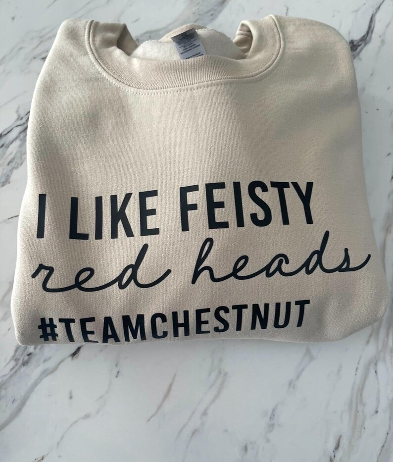 Może przedstawiać: Beżowa bluza z okrągłym dekoltem i czarnym napisem I LIKE FEISTY red heads oraz hashtagiem #TEAMCHESTNUT. Odzież posiada prążkowany kołnierz i miękką wyści&oacute;łkę wewnętrzną, zaprezentowana w złożonej formie z wyraźnym, czytelnym nadrukiem.