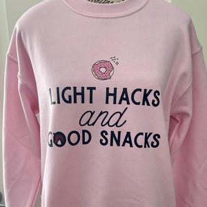 Peut inclure: Sweat-shirt rose avec un motif noir qui dit "Light Hacks and Good Snacks" avec une illustration de donut rose au-dessus du texte.