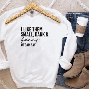 Pu&ograve; includere: Una felpa bianca a maniche lunghe con girocollo e testo nero che recita I LIKE THEM SMALL, DARK & fancy #TEAMBAY. Il capo &egrave; esposto su una gruccia con paillettes dorate, accompagnato da jeans blu, un bicchiere di caff&egrave; e stivaletti color cuoio.