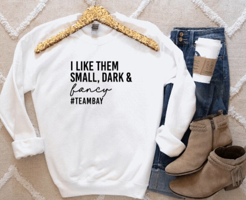 Pu&ograve; includere: Una felpa bianca a maniche lunghe con girocollo e testo nero che recita I LIKE THEM SMALL, DARK & fancy #TEAMBAY. Il capo &egrave; esposto su una gruccia con paillettes dorate, accompagnato da jeans blu, un bicchiere di caff&egrave; e stivaletti color cuoio.