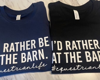 以下が含まれることがあります： ネイビーブルーとブラックの半袖クルーネックTシャツ2枚。どちらのシャツも、胸元に白い文字で「I'D RATHER BE AT THE BARN.」とプリントされており、その下に白い筆記体フォントでハッシュタグ「#equestrianlife」がデザインされています。