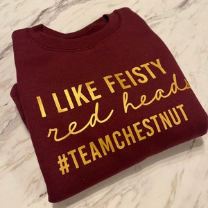Może przedstawiać: Bordowa bluza z okrągłym dekoltem i złotym napisem I LIKE FEISTY red heads #TEAMCHESTNUT. Odzież posiada prążkowany kołnierzyk i długie rękawy, a tekst jest nadrukowany na klatce piersiowej przy użyciu kombinacji czcionek blokowych i kursywy.