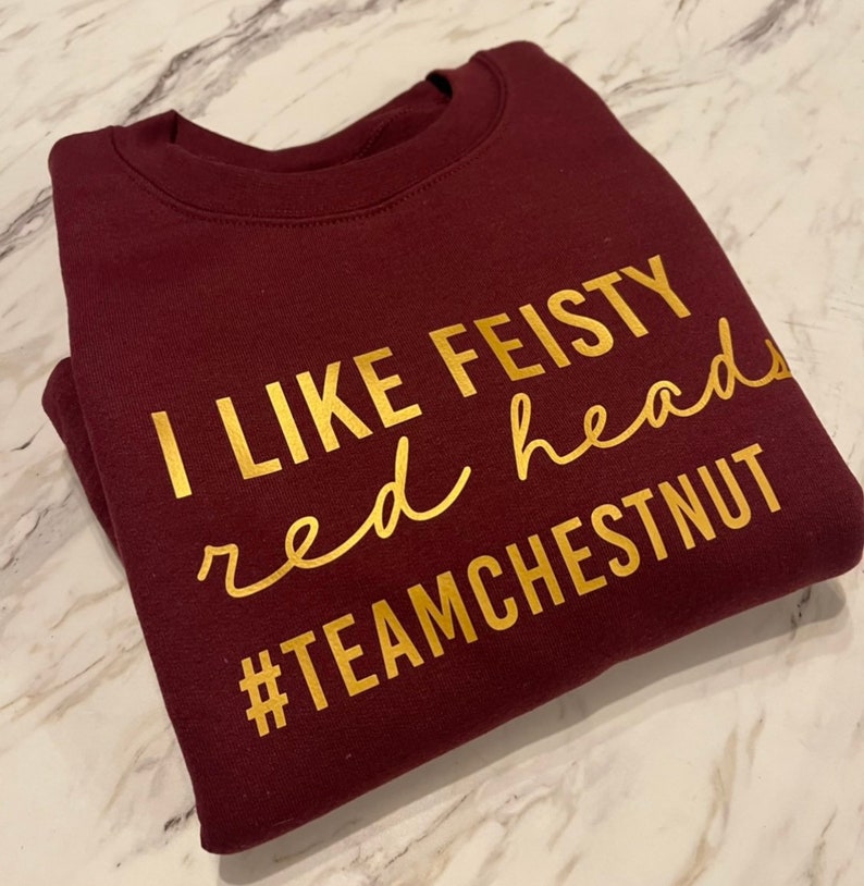 Może przedstawiać: Bordowa bluza z okrągłym dekoltem i złotym napisem I LIKE FEISTY red heads #TEAMCHESTNUT. Odzież posiada prążkowany kołnierzyk i długie rękawy, a tekst jest nadrukowany na klatce piersiowej przy użyciu kombinacji czcionek blokowych i kursywy.