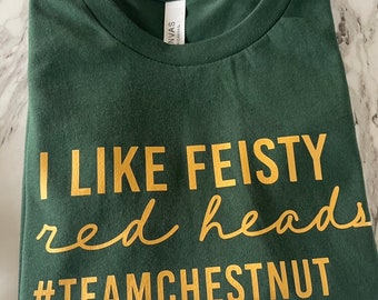 Puede incluir: Una camiseta de color verde oscuro con cuello redondo y letras doradas que dicen I LIKE FEISTY red heads #TEAMCHESTNUT. La prenda tiene un diseño estándar de manga corta con una etiqueta de marca visible en el interior del cuello.