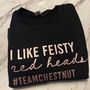 Może przedstawiać: Czarna bluza z okrągłym dekoltem i napisem w kolorze r&oacute;żowego złota o treści I LIKE FEISTY red heads oraz hashtagiem #TEAMCHESTNUT. Odzież posiada prążkowany kołnierzyk i długie rękawy, a tekst jest nadrukowany czcionką blokową oraz kursywą.