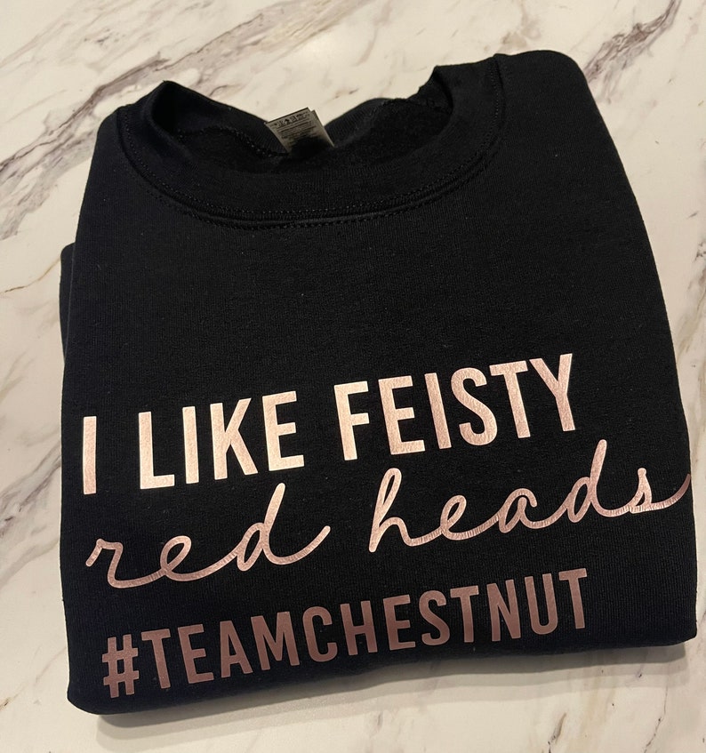 Może przedstawiać: Czarna bluza z okrągłym dekoltem i napisem w kolorze r&oacute;żowego złota o treści I LIKE FEISTY red heads oraz hashtagiem #TEAMCHESTNUT. Odzież posiada prążkowany kołnierzyk i długie rękawy, a tekst jest nadrukowany czcionką blokową oraz kursywą.