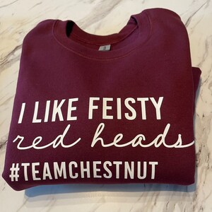 Może przedstawiać: Bordowa bluza z okrągłym dekoltem z białym napisem I LIKE FEISTY red heads oraz #TEAMCHESTNUT. Odzież posiada prążkowany kołnierzyk i długie rękawy, a napis wykonany jest w połączeniu białych liter drukowanych oraz pisma odręcznego.