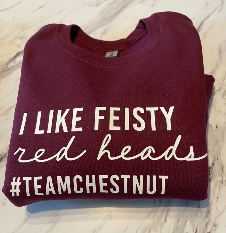Może przedstawiać: Bordowa bluza z okrągłym dekoltem z białym napisem I LIKE FEISTY red heads oraz #TEAMCHESTNUT. Odzież posiada prążkowany kołnierzyk i długie rękawy, a napis wykonany jest w połączeniu białych liter drukowanych oraz pisma odręcznego.