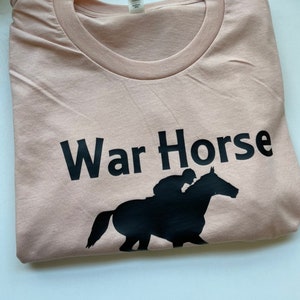 May include: A light peach-colored crew neck t-shirt featuring the black text "War Horse" printed above a black silhouette graphic of a jockey riding a horse. The garment has a standard short-sleeve design and a visible interior neck label.