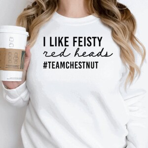 Może przedstawiać: Biała bluza z długim rękawem z czarnym napisem I LIKE FEISTY red heads #TEAMCHESTNUT. Odzież posiada okrągły dekolt i prosty kr&oacute;j, a napis wykonany jest przy użyciu kombinacji pogrubionych liter blokowych oraz czcionki imitującej pismo odręczne.