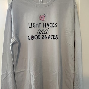 Peut inclure: T-shirt gris clair à manches longues avec l'inscription noire « LIGHT HACKS and GOOD SNACKS ». Un motif de donut rose est au-dessus du texte. Le t-shirt est suspendu à un cintre blanc.