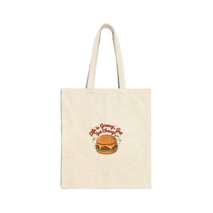 May include: Cream-colored tote bag with a burger graphic. The burger has a bun, patty, cheese, lettuce, and tomato. The text above the burger says "Life is Greasy, Get Cheesy, Get". The bag has two handles.