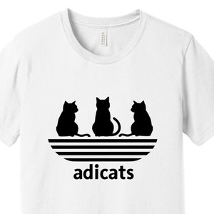 May include: White t-shirt with a black graphic of three cats sitting in a row, with the word "adicats" below them. The graphic is a parody of the Adidas logo.