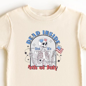 May include: Cream-colored t-shirt featuring a skeleton wearing sunglasses with a star-spangled design and holding an American flag. The shirt has the text "DEAD INSIDE But It's 4th of July" in a retro style.