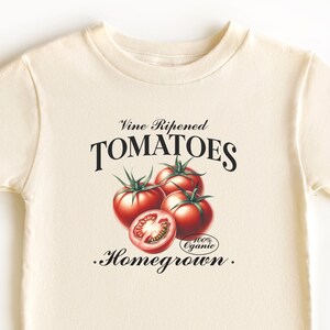 May include: A cream-colored t-shirt with a vintage-style graphic of tomatoes and the text "Vine Ripened TOMATOES .Homegrown."  The graphic includes a red and green tomato with a white label that says "100% Organic".