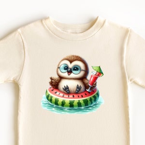 Könnte beinhalten: Ein cremefarbenes T-Shirt mit einer Cartoon-Illustration eines Eulen, das eine Sonnenbrille trägt und auf einem Wassermelonen-Schwimmring in einem Pool schwimmt. Die Eule hält ein rotes Getränk mit einem kleinen Schirm.