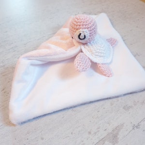 Peut inclure: Un jouet en forme de tortue en crochet rose et blanc attaché à une couverture pour bébé blanche et rose.