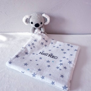 Puede incluir: Un juguete de koala de ganchillo gris con orejas blancas y esponjosas se encuentra sobre una manta de bebé blanca y gris con estampado de estrellas. El nombre "MATHEO" está bordado en negro.