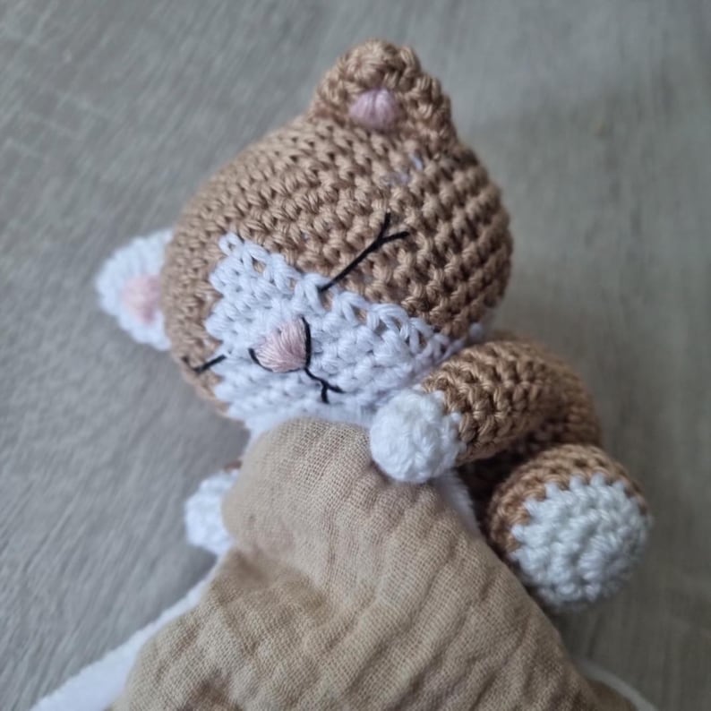 May include: A crocheted cat security blanket featuring a tan and white yarn head with embroidered closed eyes and a pink nose. The cat has white paws and is attached to a soft, tan-colored muslin fabric square that provides a textured, layered appearance.