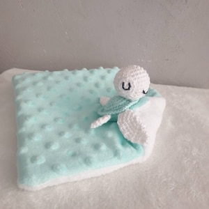 Peut inclure: Une couverture pour bébé avec un tissu à pois minky turquoise et un envers en peluche blanche. Une tortue crochetée avec une tête blanche, une carapace turquoise et des nageoires blanches est attachée. La tortue a des yeux brodés noirs.